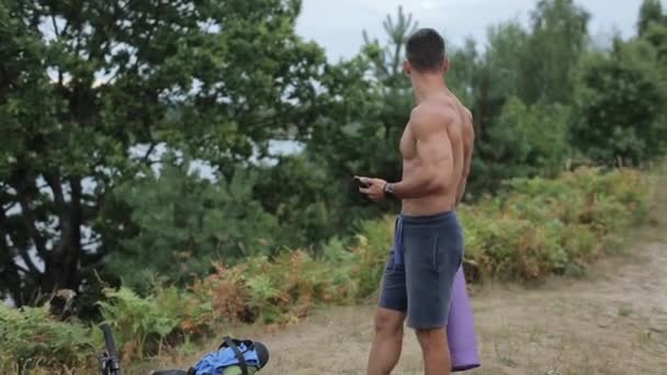 Молодий м'язистий чоловік з телефоном і спортивним килимком у руках прогулюється вздовж берега річки, щоб вибрати місце для тренувань. Крупним планом. Вид збоку — стокове відео