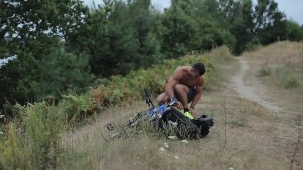 Um jovem homem musculoso de calções azuis agachamentos ao lado de sua bicicleta e mochila e amarra seus tênis antes de treinar na natureza, em seguida, levanta-se com um telefone na mão. Vista frontal. Close-up — Vídeo de Stock