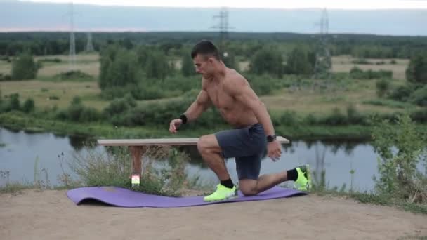 Młody muskularny mężczyzna z nagim tułowiem ćwiczy nad rzeką i huśta się nogą wspinając się na ławkę. Widok z boku. Zbliżenie. Kamera porusza się w górę i w dół — Wideo stockowe