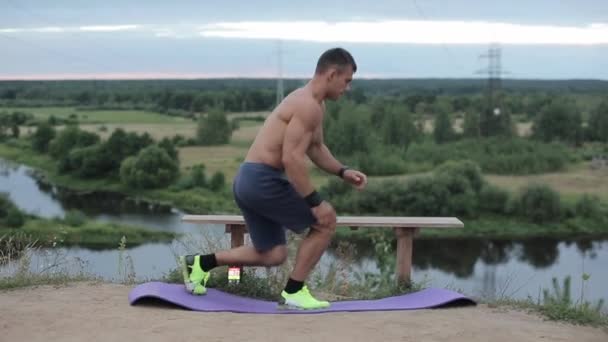 Ung kroppsbyggare i blå shorts tåg vid floden och gör steg övningar sätta foten på en träbänk. Sidovy. Närbild. Kameran rör sig bakom honom. — Stockvideo