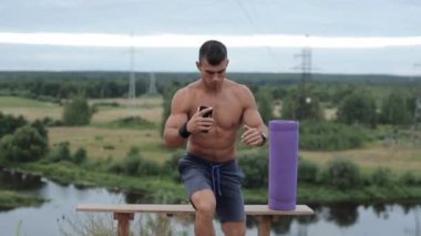 Karın kasları olan genç bir vücut geliştirici nehir kıyısında egzersiz yapıyor. Tek ayağı üzerinde çömeliyor ve elinde bir telefon tutuyor. Ön manzara. Yakın plan.