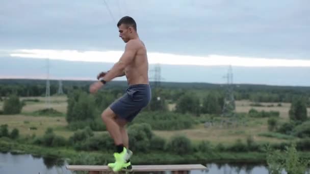 A young muscular man with a naked torso works out by the river and does leg swings as he climbs onto a bench. Side view. Close-up. Camera moves up and down — Stock Video