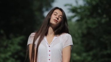 Gülümseyen genç bir kız, arka planda duran yeşil ağaçların önünde uzun saçlarını eliyle düzleştiriyor. Ön manzara. Yakın plan. Bulanık arkaplan