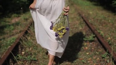 Uzun beyaz elbiseli genç bir kız ormanda terk edilmiş bir demiryolu boyunca çıplak ayakla yürüyor.