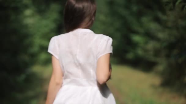 一个穿着白衣的小女孩赤着脚沿着森林里一条废弃的旧铁路的铁轨跑着，用手把她的长发拉直。后视镜慢动作 — 图库视频影像