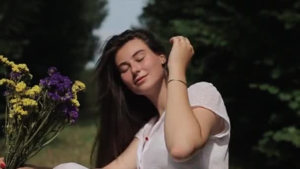 Něžná mladá žena v bílých šatech sedí na pozadí přírody s kyticí divokých květin, očichává je a zavírá oči. Detailní záběr. Zpomal. Boční pohled — Stock video