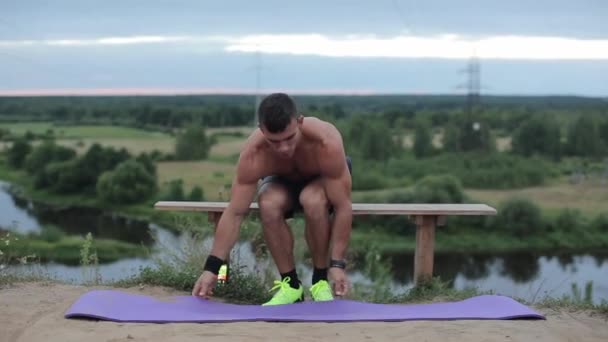 Young muscular guy with a naked torso goes in for outdoor sports by the river and straightens a sports mat before exercising. Close-up — Stock Video