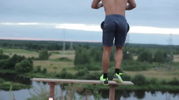 Mladý svalnatý muž v modrých trenýrkách dělá kroková cvičení u řeky skákající na lavičku. Pohled zezadu. Detailní záběr. Kamera se pohybuje kolem objektu a nahoru a dolů — Stock video