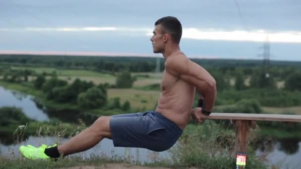 Un jeune bodybuilder sans t-shirt va faire du sport dans la nature et secoue ses muscles des bras en faisant des pompes du banc. Vue latérale. Gros plan. La caméra se déplace de haut en bas — Video