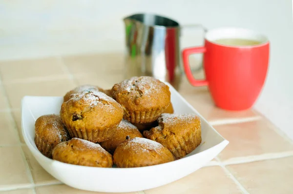 Muffins con azúcar en polvo en un plato y una jarra de leche con una taza . — Foto de Stock
