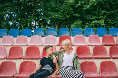 beautiful mother and son looking each other while sitting together on stadium seats clipart