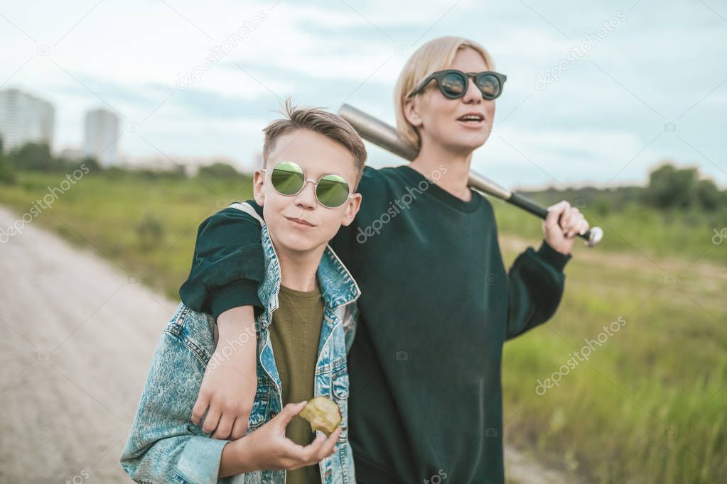 mother and son in sunglasses walking together on ground road, woman holding baseball bat and boy eating apple 