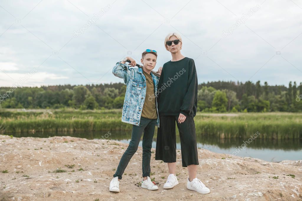 mother and son in sunglasses standing with baseball bat and looking at camera near lake