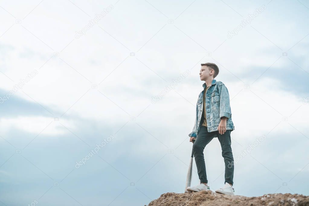 low angle view of boy in denim jacket standing with baseball bat and looking away at cloudy day