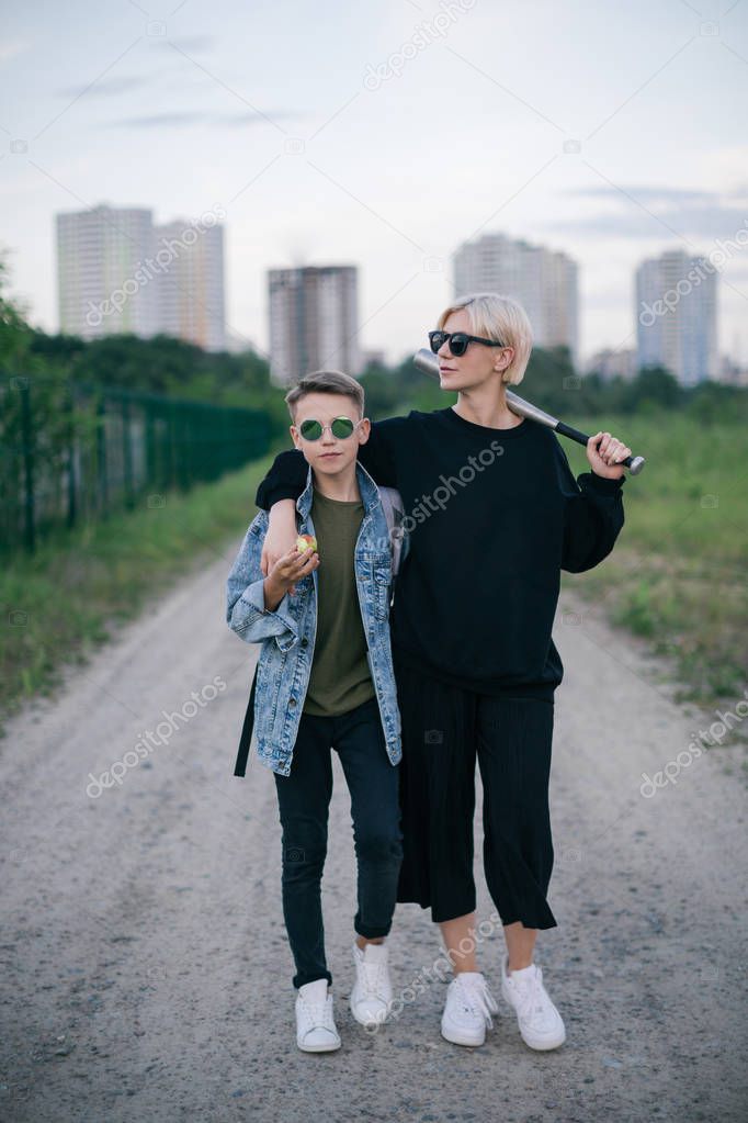 stylish mother and son in sunglasses standing with baseball bat on path