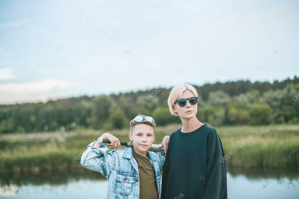 beautiful stylish mother and son standing with baseball bat near lake 