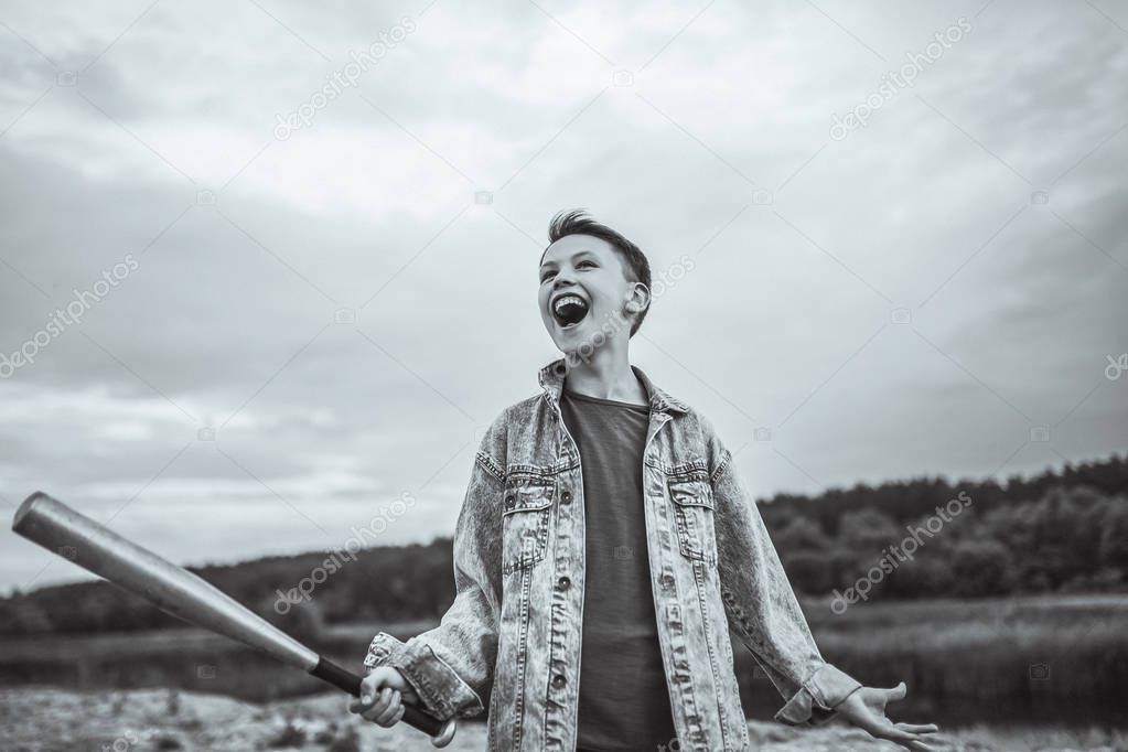 low angle view of excited boy in denim jacket holding baseball bat outdoors, black and white photo