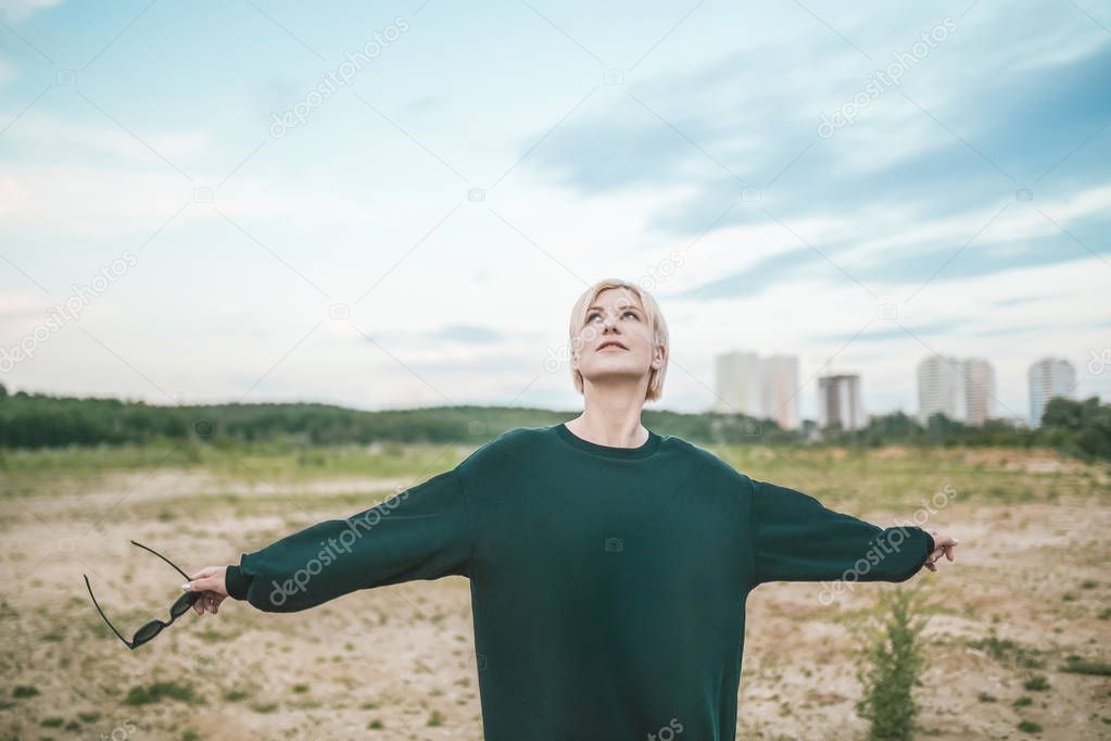 beautiful blonde woman holding sunglasses and looking up while standing with open arms at cloudy day