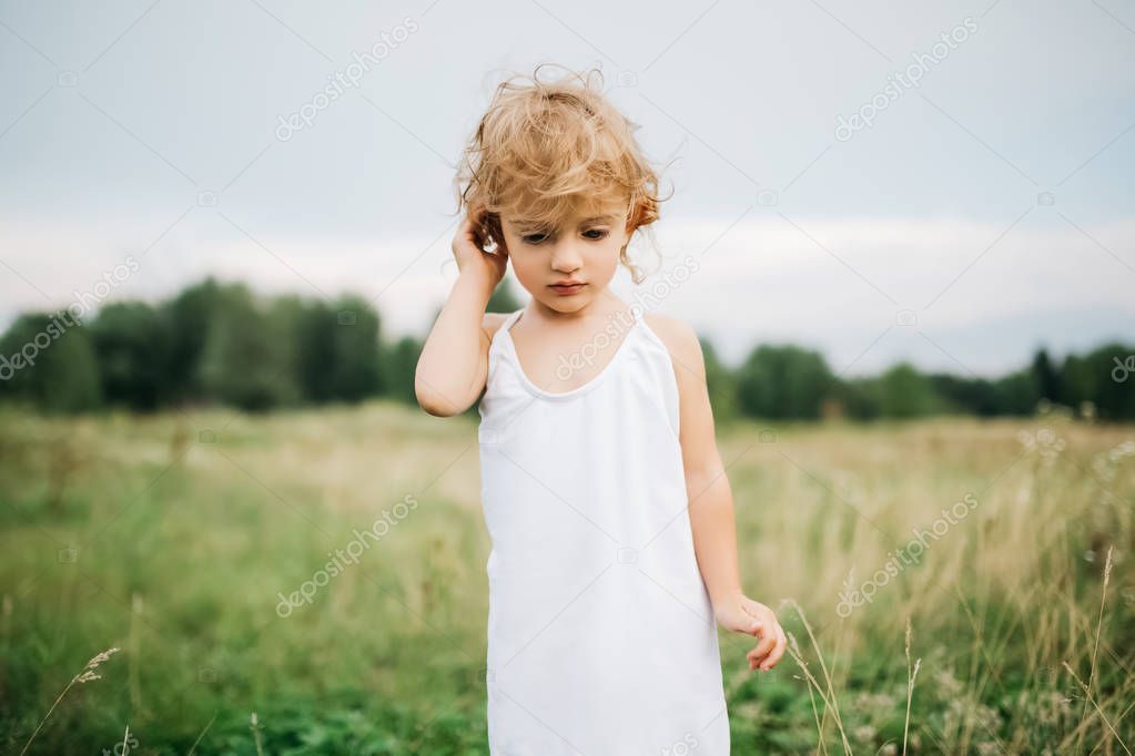 adorable child with curly hair standing in field and looking down