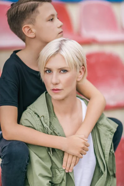 Beautiful preteen boy hugging mother while sitting together on stadium — Stock Photo