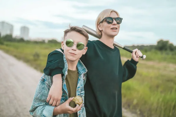 Madre e hijo en gafas de sol caminando juntos en el camino de tierra, mujer sosteniendo bate de béisbol y niño comiendo manzana - foto de stock