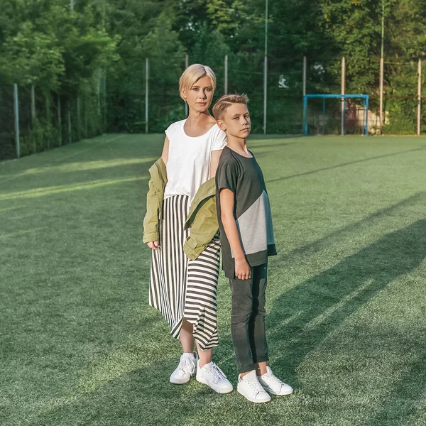 Full length view of beautiful stylish mother and son looking at camera while standing together in green lawn — Stock Photo