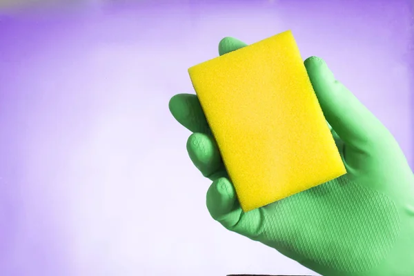 Hand in green glove holding a yellow sponge on a blue background