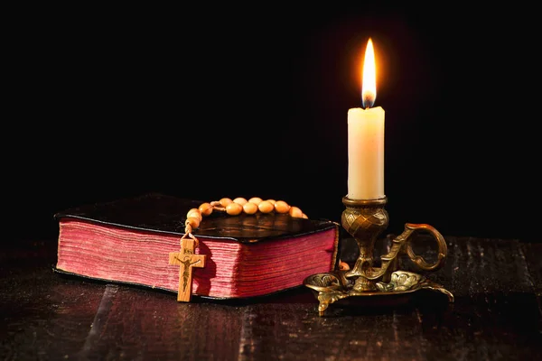 Rosary on the book, and a lit candle in a candlestick on a black background