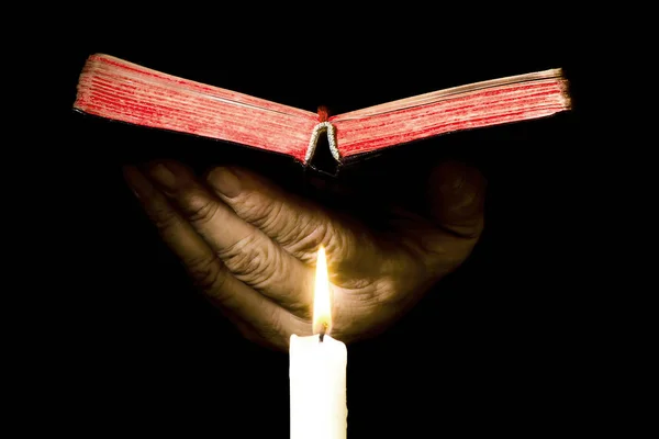 Handen håller en öppen bok över ett tänt ljus, isolerad på en svart Stockbild