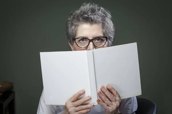 Stylish elderly woman hiding behind a book looking at camera. White cover with empty space for text.