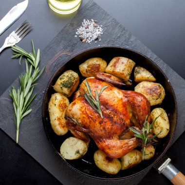 Fırında kızartılmış tavuk, patates dilimleri ve siyah arka planda dökme demir tavada taze biberiye. Yukarıdan görünüm