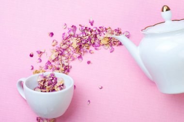 Kuru bitki çayı, beyaz bir porselen çaydanlıktan pembe bir yüzey üzerinde bir fincana dökülen bir çay gülüdür. Yukarıdan bak. Çay partisi konsepti.