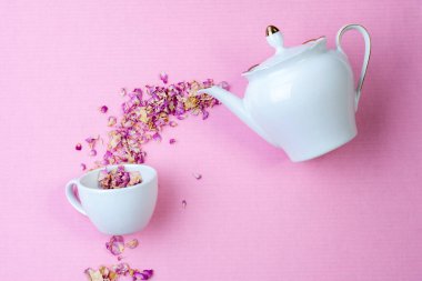 Kuru bitki çayı, beyaz bir porselen çaydanlıktan pembe bir yüzey üzerinde bir fincana dökülen bir çay gülüdür. Yukarıdan bak. Çay partisi konsepti.