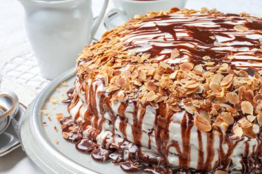 Beyaz katmanlı badem keki, erimiş çikolatayla birlikte porselen seti, klasik çatallar, badem taneleri olan beyaz masa örtüsü.