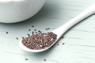 Beyaz porselen kâse ve kaşıkta açık mavi arka planda izole edilmiş Chia tohumları (Salvia hispanica). Sağlıklı yiyecekler, abur cubur ve takviye konsepti. Yukarıdan bak. Düz yatıyordu.