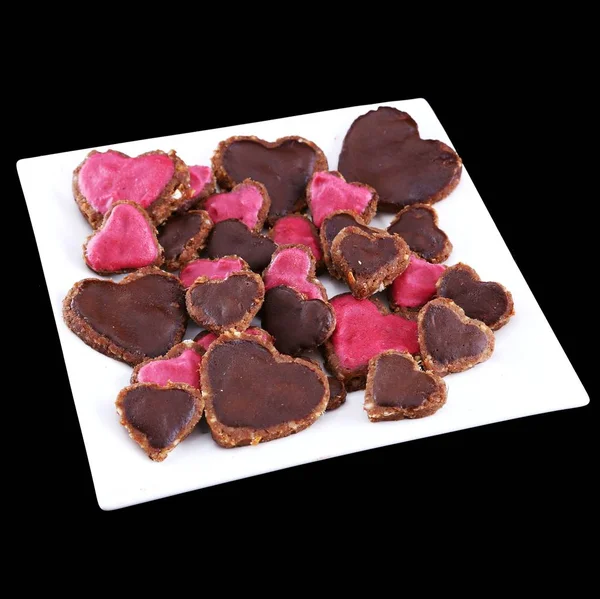 Vegan raw-food heart-shaped cookies served on a square plate isolated on black background