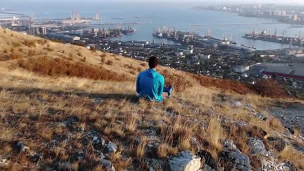 Ein junger Mann sitzt auf einem Berg und demonstriert den Zoomeffekt. Schießen von hinten — Stockvideo