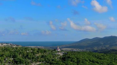 Yeşil alanlar, orman, koy, deniz, dağlar, mezarlık ve arka planda evleri olan dağın tepesindeki bir kilisenin panoramik görüntüsü.