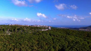 Yeşil alanlar, orman, koy, deniz, dağlar, mezarlık ve arka planda evleri olan dağın tepesindeki bir kilisenin panoramik görüntüsü.