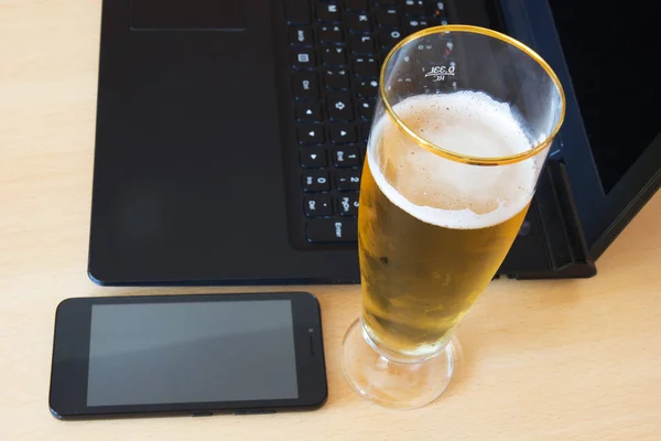 Glass of beer, laptop computer and smartphone on table.