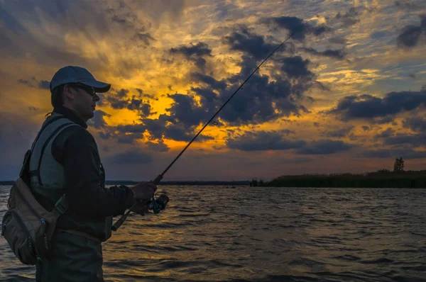 Silhouette des Fischers. Mann im Wasser mit Angelrute vor wolkenverhangenem Sonnenuntergang — Stockfoto