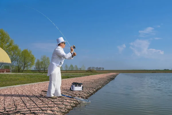 Изысканная рыбалка. Рыбак в белом костюме ловит рыбу, крутя удочку на озере. — стоковое фото