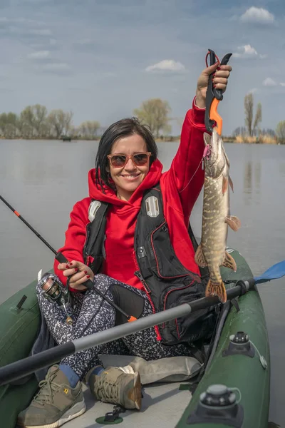 Kayak fishing. Fisher girl holding pike fish trophy on inflatable boat with fishing tackle at lake.