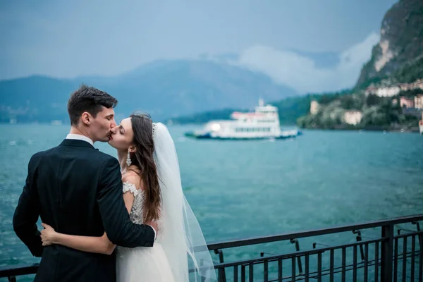 在多云的天气里 一对婚礼夫妇在湖和山的背景下接吻 — 图库照片