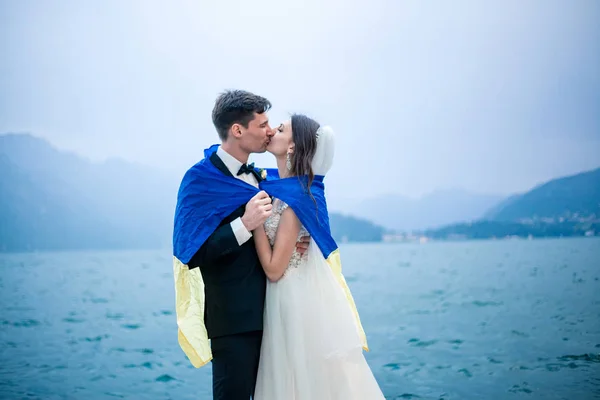 在多云的天气里 一对婚礼夫妇在湖和山的背景下接吻 — 图库照片