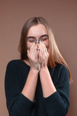 Kız gülüyor ve bej renkli arka plan üzerinde siyah süveterli elleriyle yüzünü kapsar