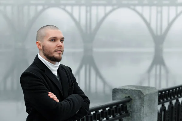 short-haired young man in a black coat looks into the distance on the embankment against the background of the bridge. fog