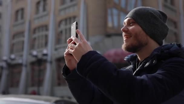 Um jovem de casaco azul e boné cinza atira em uma cidade pelo telefone — Vídeo de Stock