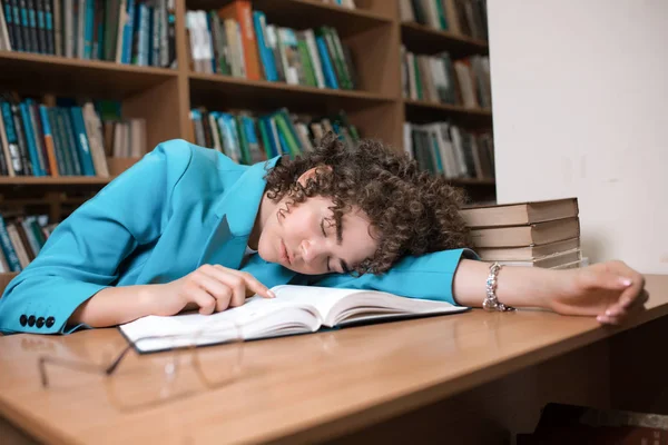 Joven chica rizada hermosa en gafas y traje azul sentado con libros en la biblioteca . —  Fotos de Stock