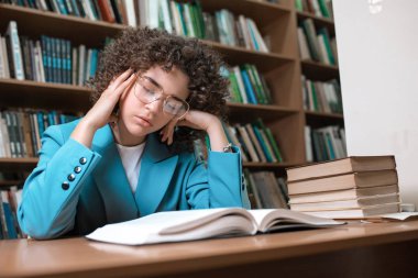 Gözlük ve mavi takım elbise li genç güzel kıvırcık kız kütüphanede kitap ile oturan.